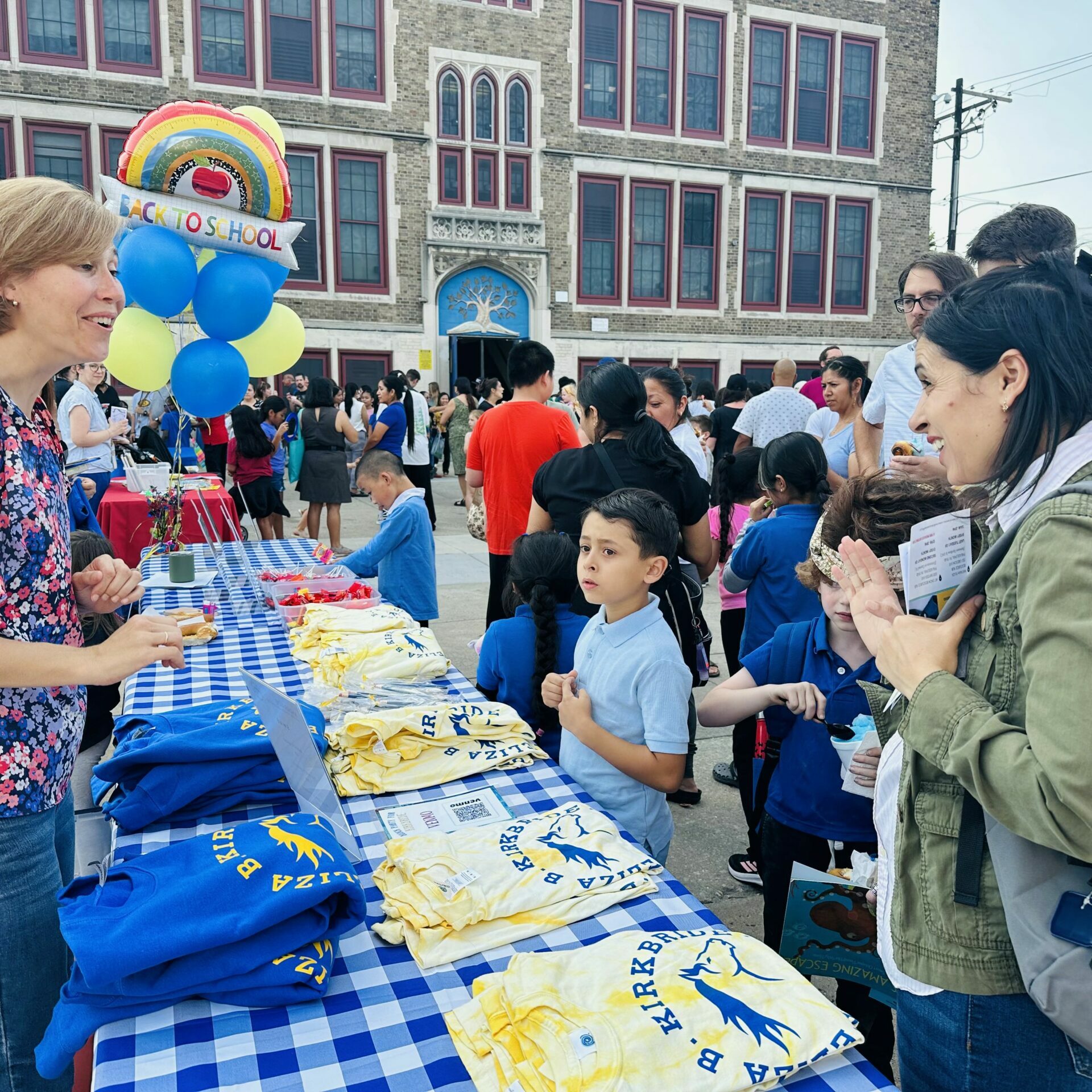 Kirkbride Elementary Back-to-School Night Families gather outside Kirkbride Elementary School during a back-to-school event. A woman greets parents at a blue-and-white checkered table displaying Kirkbride T-shirts, while balloons with a “Back to School” sign and the school building appear in the background.