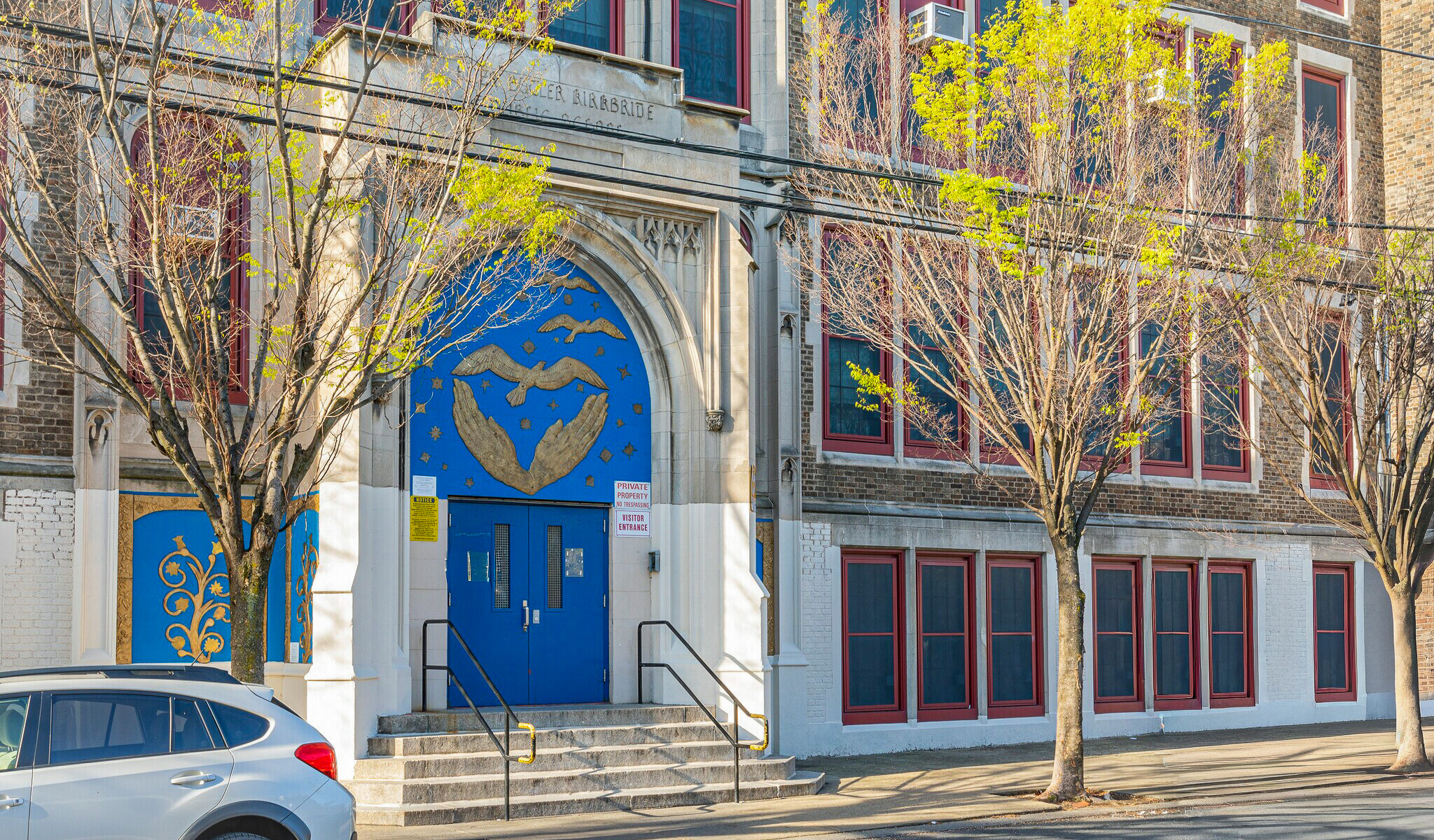 Kirkbride Elementary School Main Entrance – Philadelphia, PA Front entrance of Kirkbride Elementary School in South Philadelphia, featuring blue double doors decorated with gold artwork of hands and a dove, surrounded by arched stone detailing and red-trimmed windows with trees lining the sidewalk.